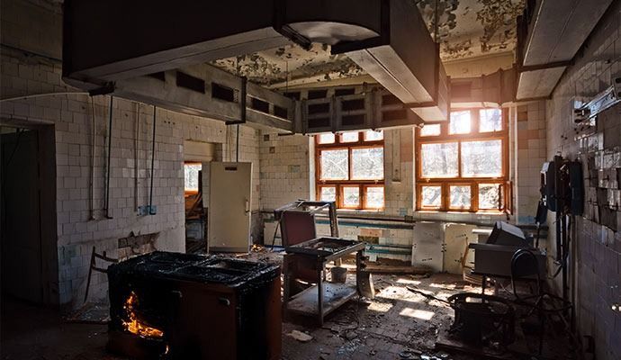 Abandoned, decaying kitchen: ovens, ventilation hood, crumbling walls, sunlit windows.