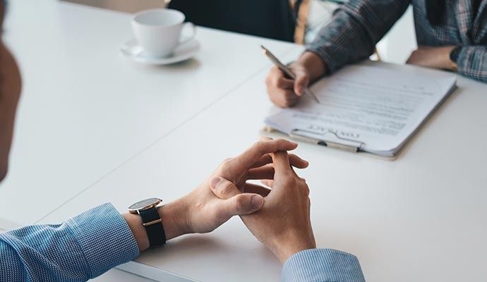 Two people at a white table, one writing on a document, the other with hands clasped, wearing a watch.