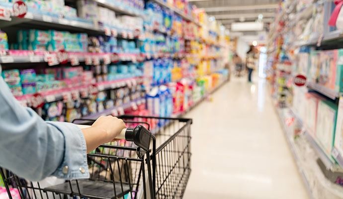 Hand pushing a shopping cart down a brightly lit supermarket aisle.