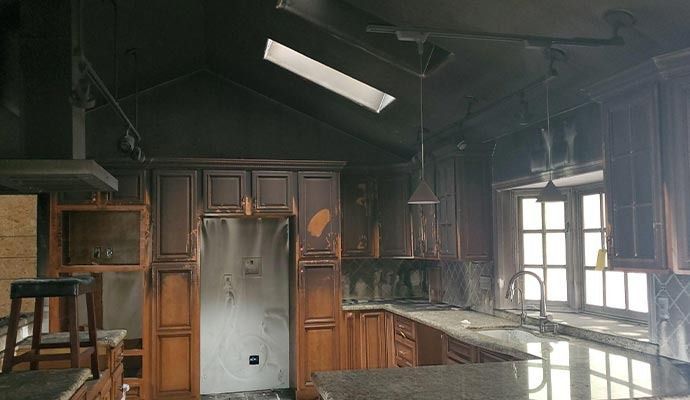 Fire-damaged kitchen. Darkened cabinets, charred walls, skylight, and ruined appliances are visible.