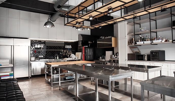 Spacious commercial kitchen with stainless steel tables, white cabinets, and black accents under a metal grid ceiling.