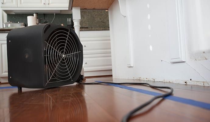 Black fan on wood floor in a kitchen, a blue tape outline, and white cabinets in the background.