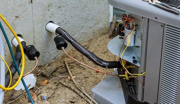 An AC unit with insulated pipes connected to a wall, electrical wiring visible, outdoors.