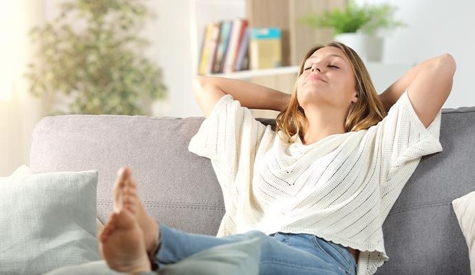 Woman relaxing on a grey couch indoors with her hands behind her head and eyes closed.