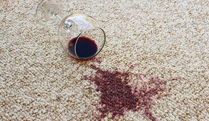 A spilled glass of red wine on a cream-colored carpet, creating a dark red stain.