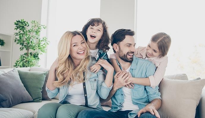 Family of four smiling on a couch; two girls playfully hug their parents.