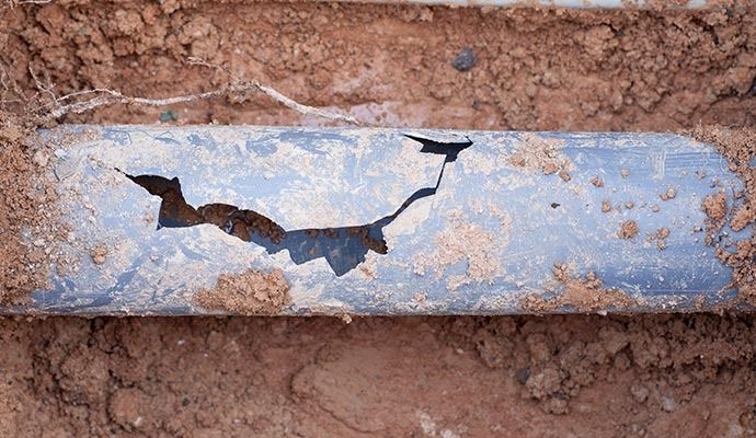 Cracked blue pipe buried in reddish-brown soil, showing significant damage.