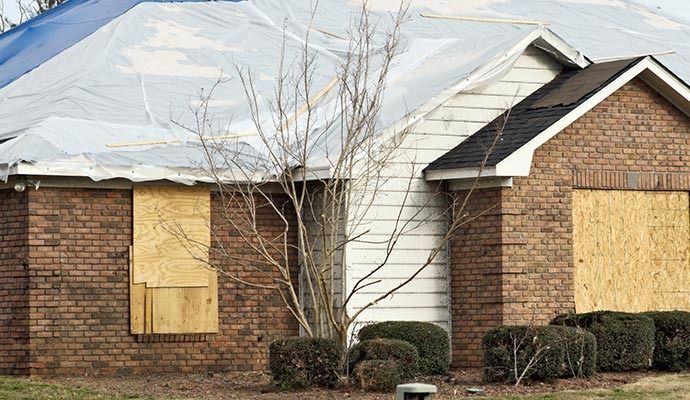 House with boarded-up windows, covered roof, and damaged brick exterior. A small tree is in the foreground.
