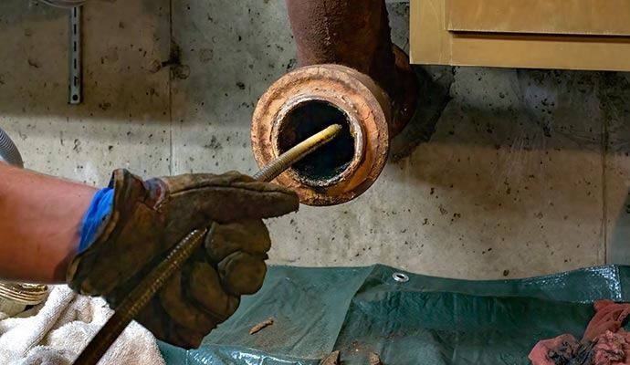 A gloved hand inserts a metal rod into a rusty pipe opening in a basement setting.
