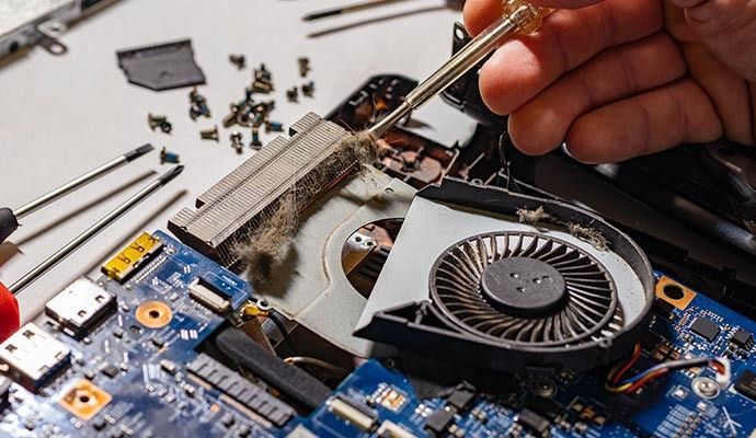 Laptop being disassembled, a hand uses a screwdriver to clean dust from the fan.