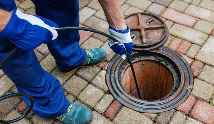 Person in blue coveralls and gloves inspecting a sewer access point with a black cable outdoors.
