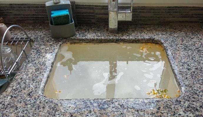 Kitchen sink filled with dirty, stagnant water and debris. Granite countertop, faucet, and sponge holder visible.
