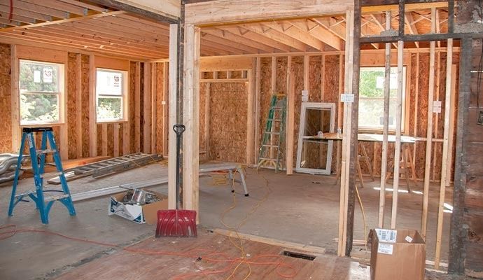 Interior view of a house under construction; wooden studs and framing visible. Light blue ladder and tools on the floor.
