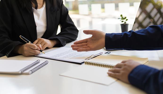 Person signing document, another person's hand extended; setting appears to be an office.