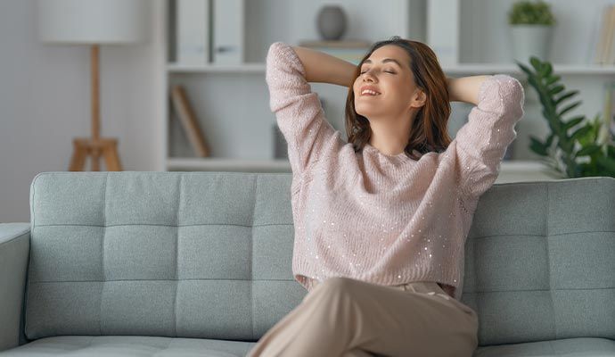 Woman relaxing on a gray sofa, hands behind head, eyes closed. Neutral setting with shelves and plants.