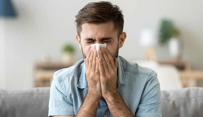 Man sneezing into a tissue, eyes closed, sitting on a couch.