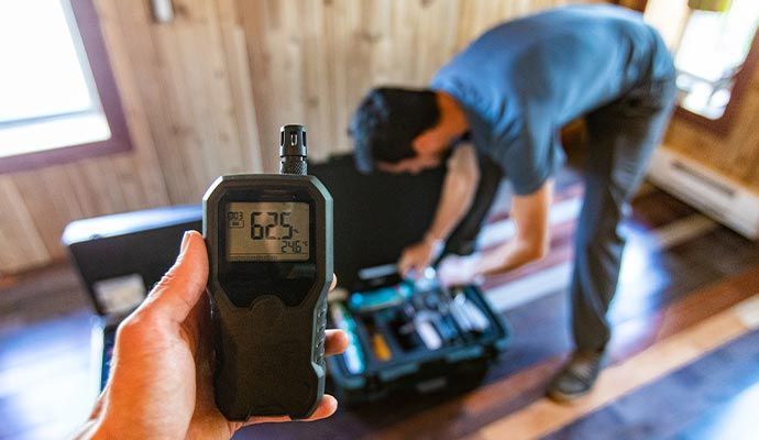 Hand holding a black meter displaying 62.5, a person working on equipment with a toolbox inside a building.