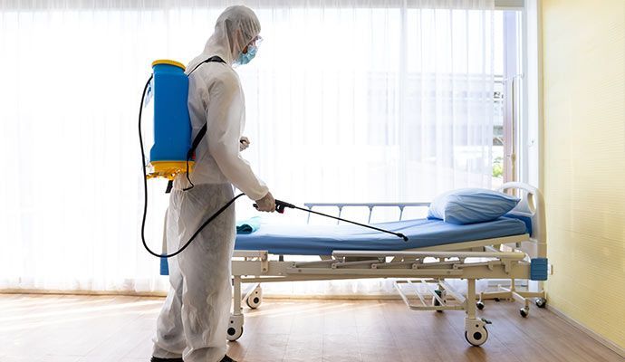 Person in hazmat suit disinfecting a hospital bed with a sprayer.