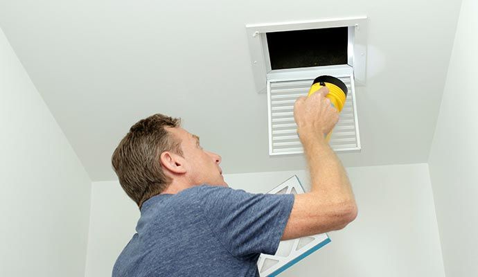 Man changing air filter in ceiling vent. He holds a yellow flashlight and filter in a white room.