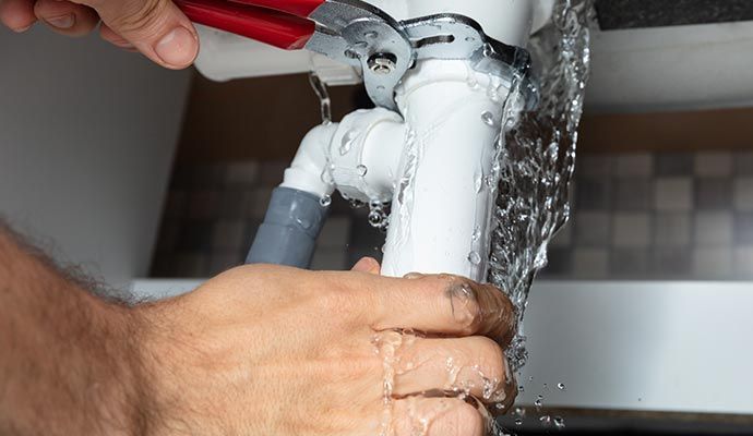 Hands with pliers tightening a pipe under a sink, water gushing out.