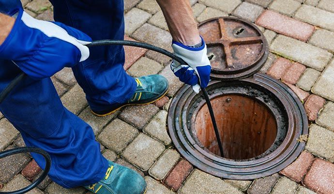 Person in blue coveralls and gloves using a tool to inspect an open sewer manhole in a brick-paved area.