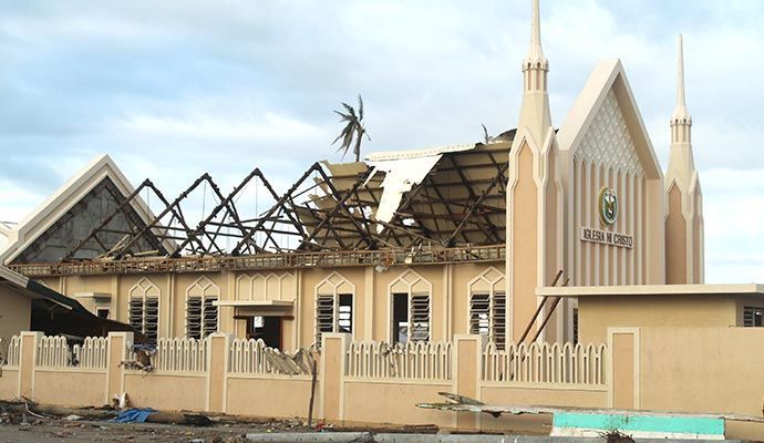 Damaged church building with roof partially collapsed; tan exterior, fence, cloudy sky.