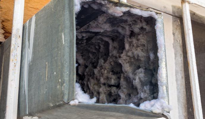 Cut-open rectangular metal duct filled with thick, gray, dusty debris and white insulation.