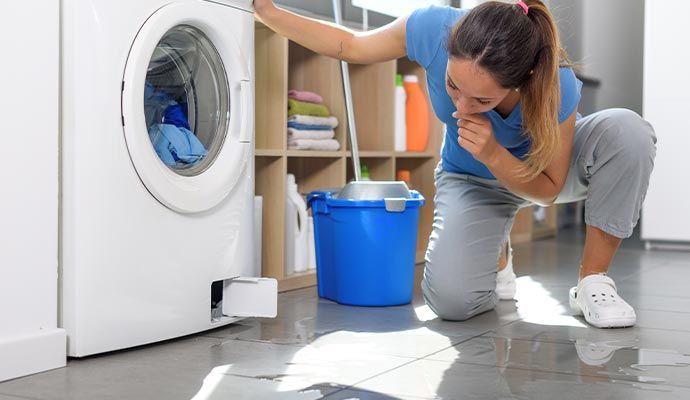 Woman kneels next to a washing machine that is leaking water. A blue bucket sits nearby.