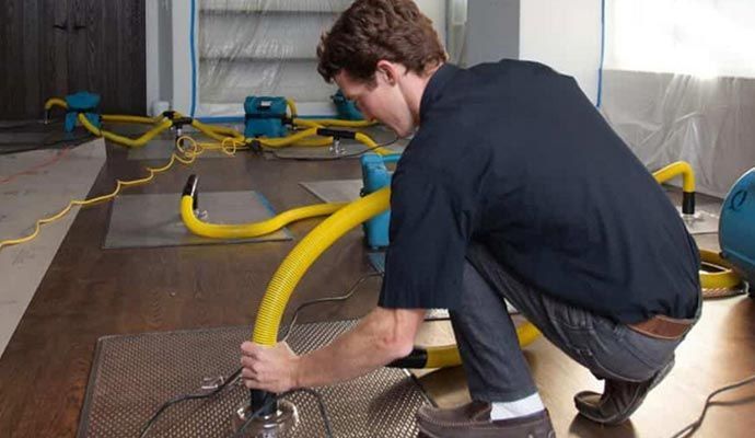 Man in black shirt installing water damage restoration equipment on a wooden floor.