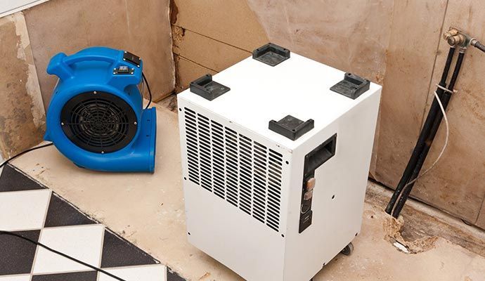 Blue air mover and white dehumidifier in a room with black and white tiled floor and damaged walls.