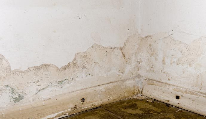 Corner of a room with water damage on the walls and floor, causing peeling paint and discoloration.