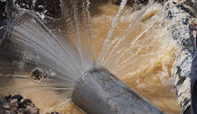 Water gushes from a broken pipe, spraying outward. Brown, muddy water surrounds it.