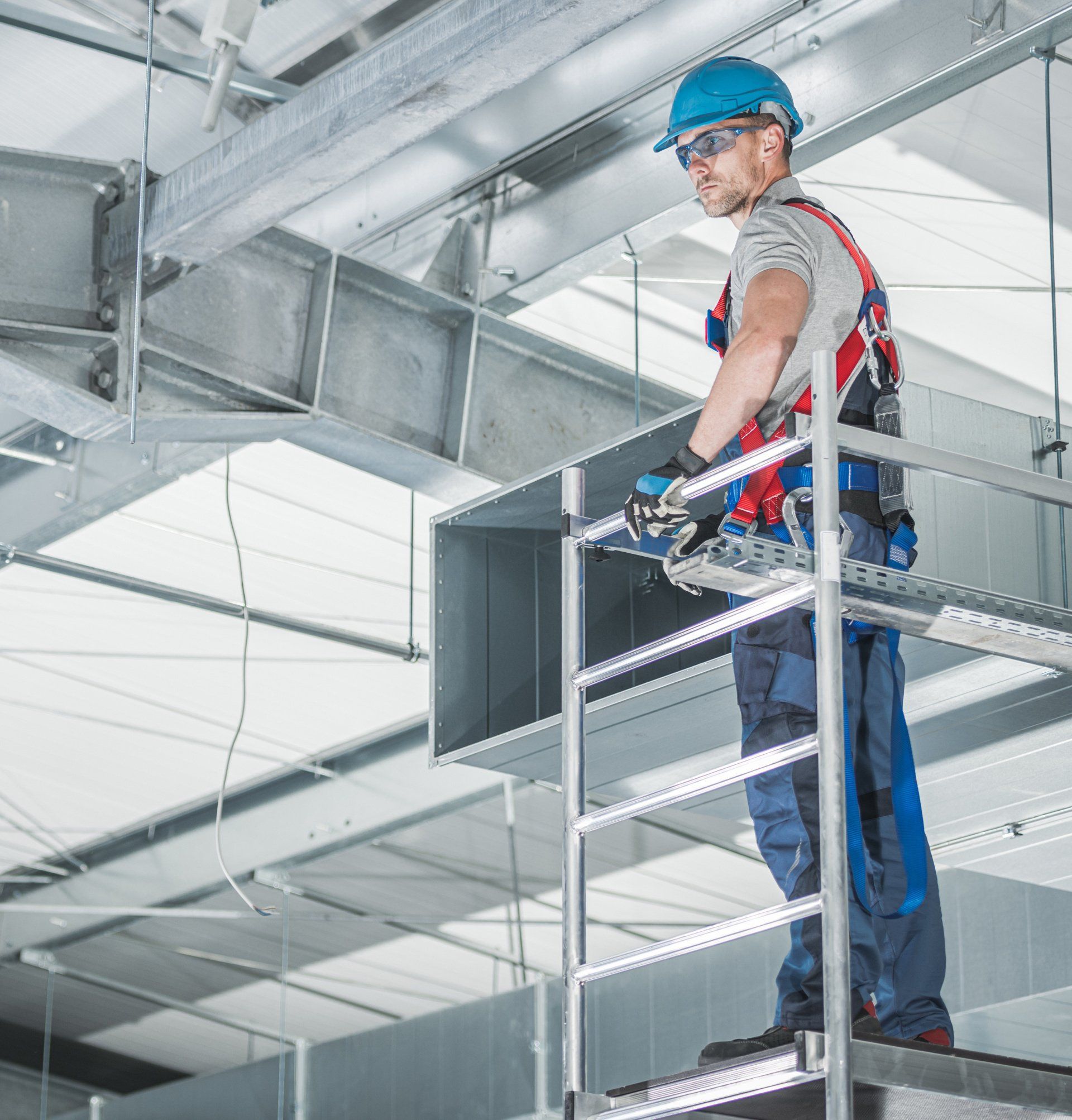 a man wearing a blue hard hat stands on a ladder repairing an HVAC system