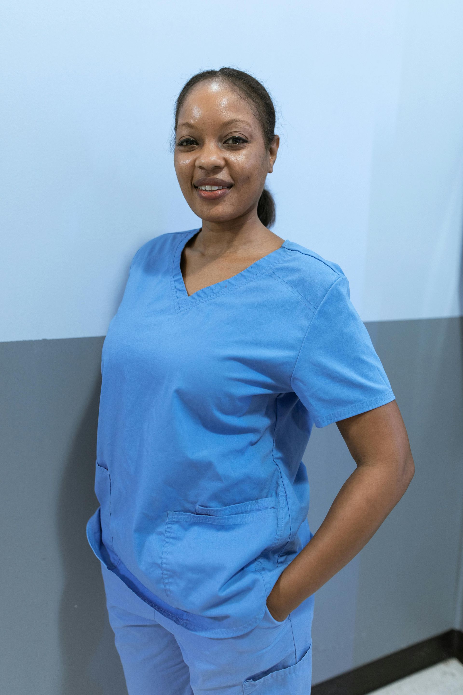 A Nurse in blue scrubs is standing in a hallway with her hands in her pockets.