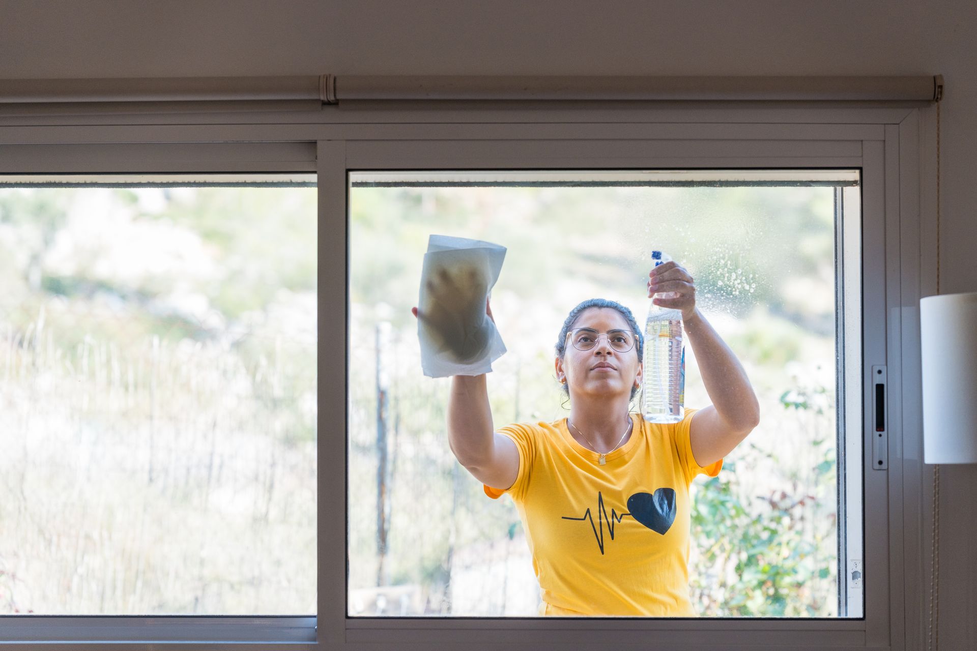 A beautiful young woman cleaning and wiping a window with a spray bottle and blotting paper A beautiful young woman cleaning and wiping a window with a spray bottle and blotting paper