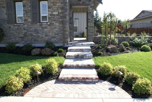 A landscaped front yard with a stone pathway, green lawn, trimmed shrubs, and a stone house entrance with a gated door.