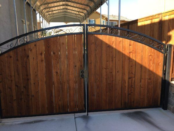 A wooden gate with decorative wrought iron accents, set in a residential driveway with a covered carport in the background.