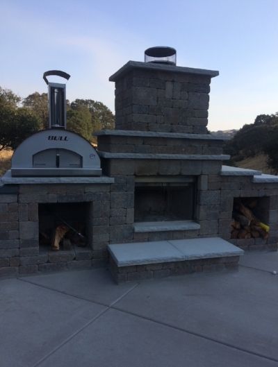 An outdoor kitchen with a brick fireplace with a pizza oven next to it.