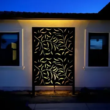 A decorative metal privacy screen with leaf patterns, illuminated by warm lighting, set against a modern home's exterior.