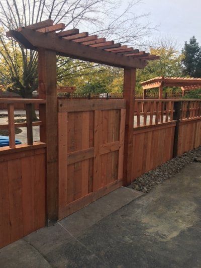 A wooden garden gate with a pergola top, set within a matching fence, surrounded by trees and a gravel pathway.