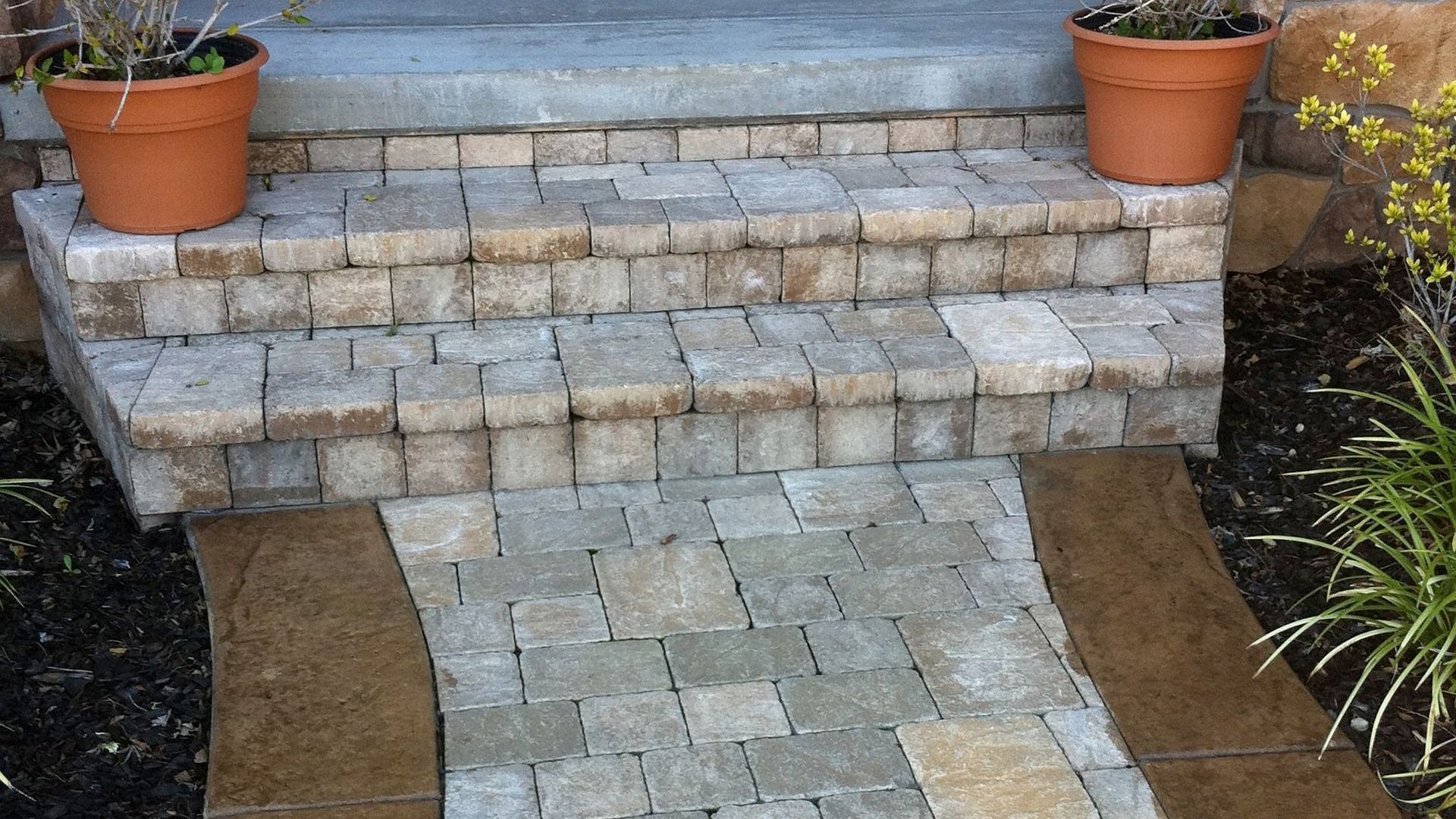 A brick walkway leading to a house with potted plants on the steps by Turfworks Inc. A paver contractor in Vacaville.