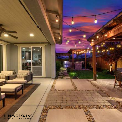 Backyard patio by Turfworks Inc featuring seating, string lights, pergola, paver paths, and an outdoor kitchen at sunset.
