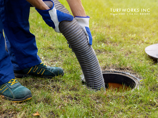 Person in blue jumpsuit and gloves inserting hose into a septic tank in grassy yard.