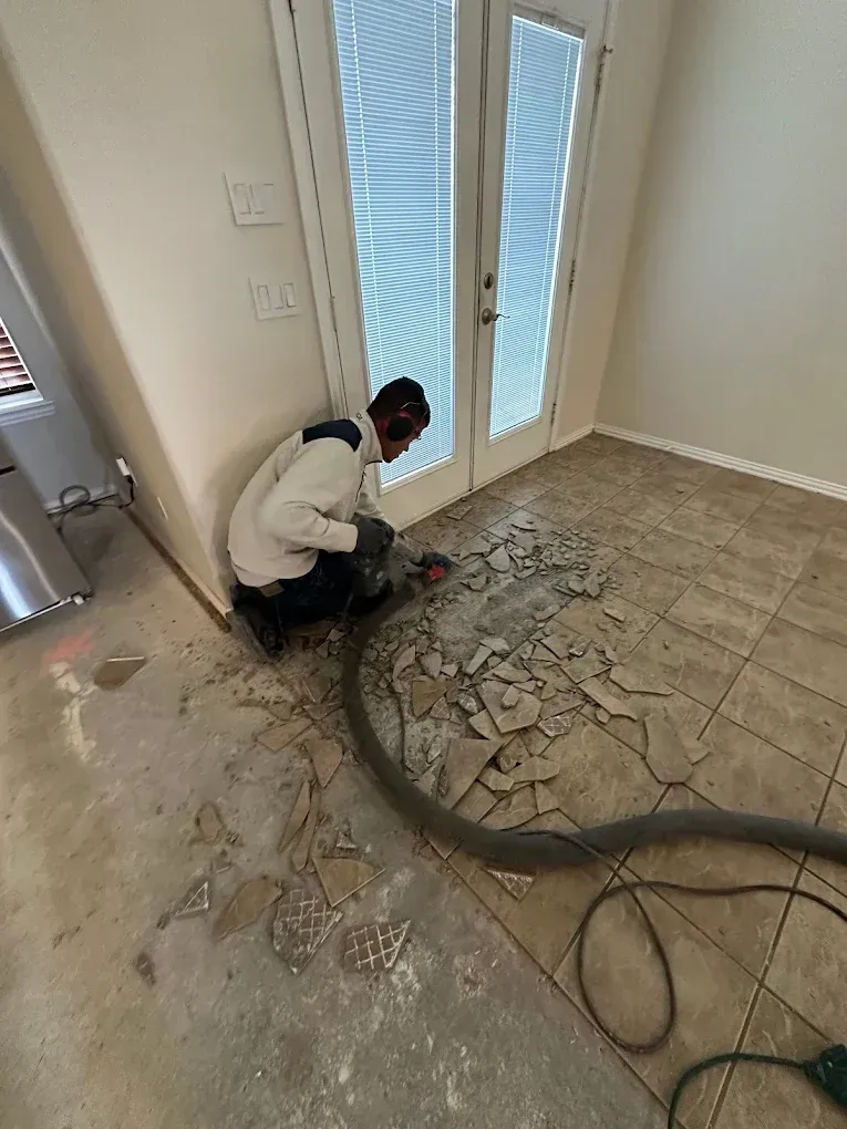 Person removing tile flooring near a door. Gray dust and debris covers the floor.