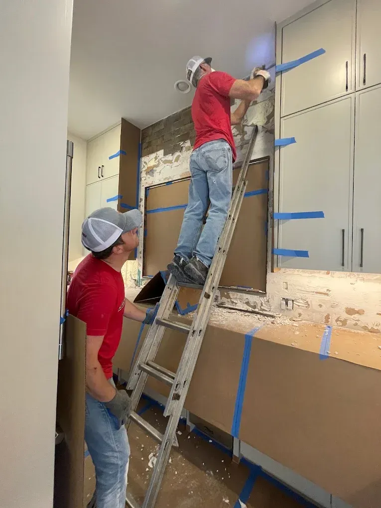 Two people in a kitchen installing something. One on a ladder using a drill, the other looking on.