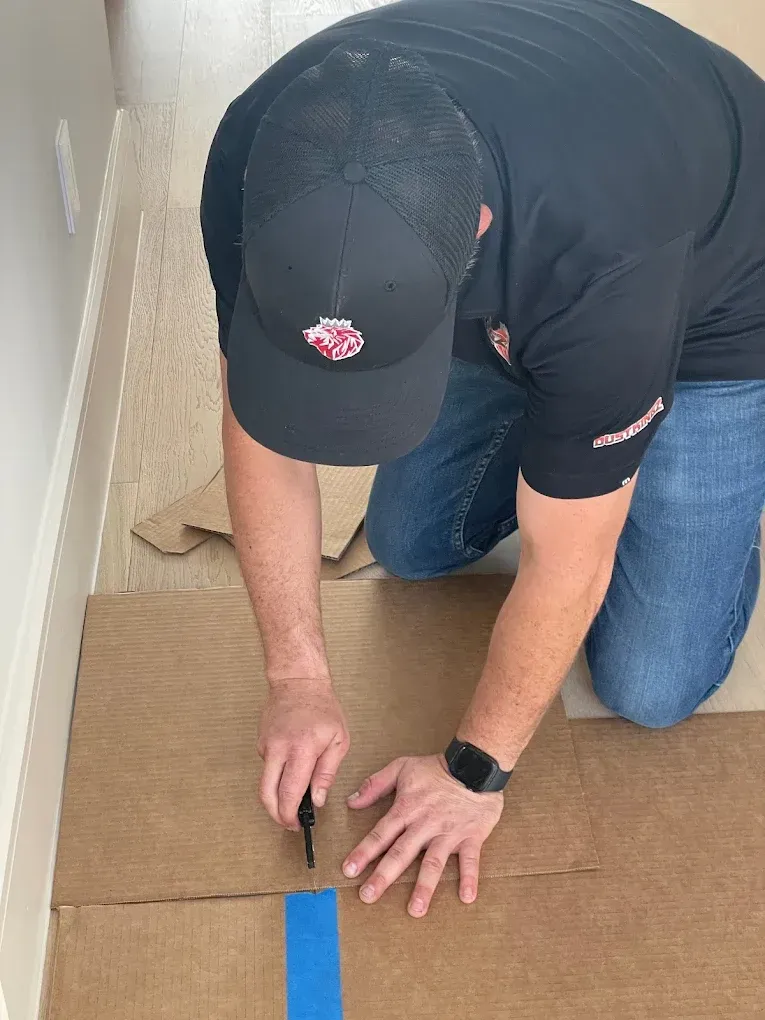 Person kneeling, cutting cardboard with a utility knife. Wearing a black cap and shirt, blue jeans.