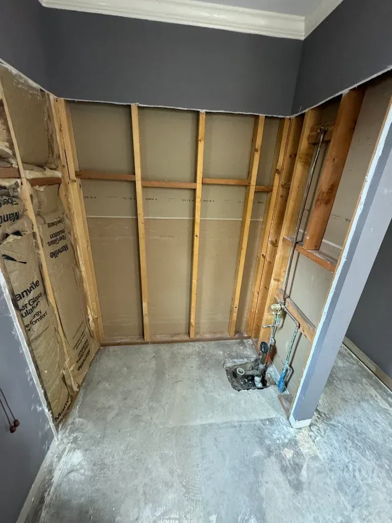Interior construction site, exposed wall studs, insulation, plumbing, and concrete floor. Grey walls and ceiling.