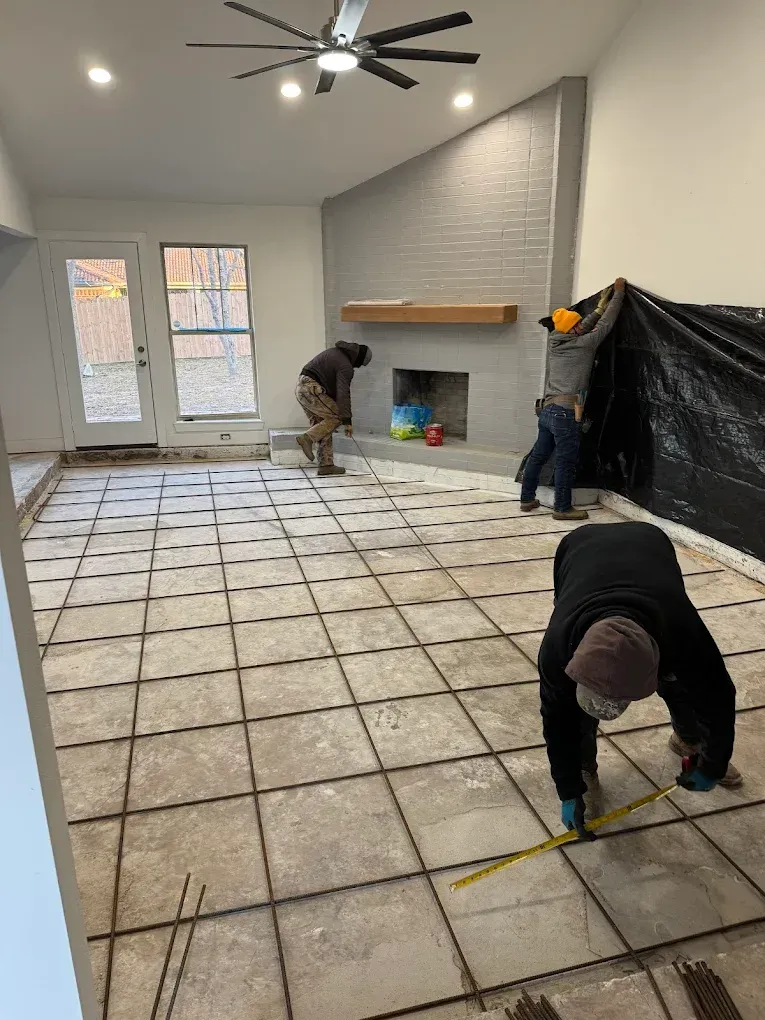 Workers installing a tile floor in a living room, measuring and covering the work area near a fireplace.