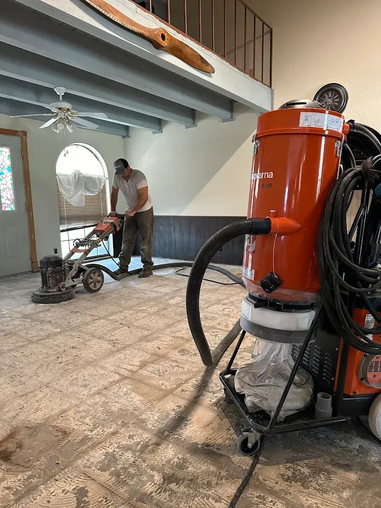 Man operating a floor grinder, dust collection system in use. Interior space with beams, an airplane propeller.