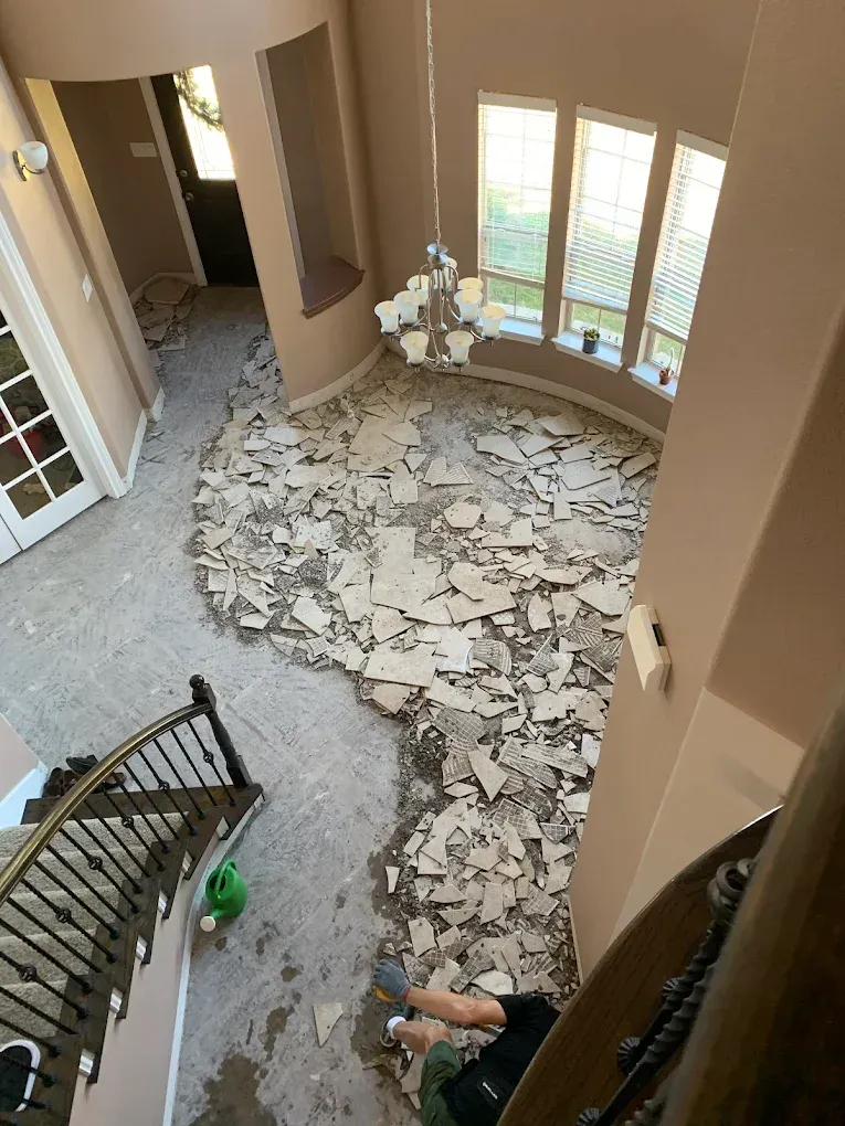 Interior view of a home undergoing floor tile removal. Debris litters the floor; a chandelier hangs.